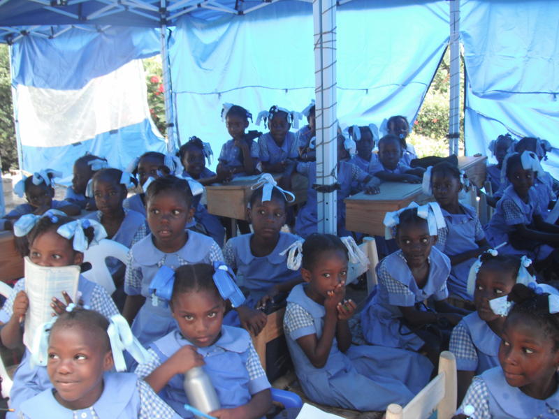 A classroom of girls in blue uniforms seated under a relief tent